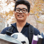 Image of a happy male-presenting student with a backpack and carrying a box of housewares, books, and a soccer ball.