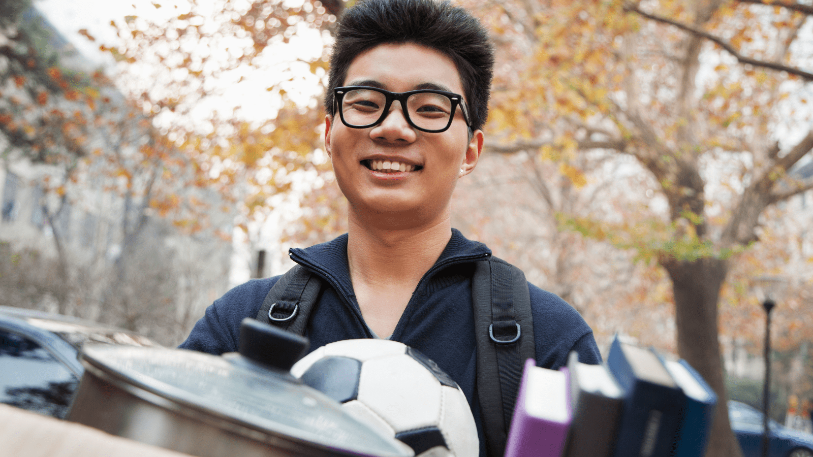 Image of a happy male-presenting student with a backpack and carrying a box of housewares, books, and a soccer ball.