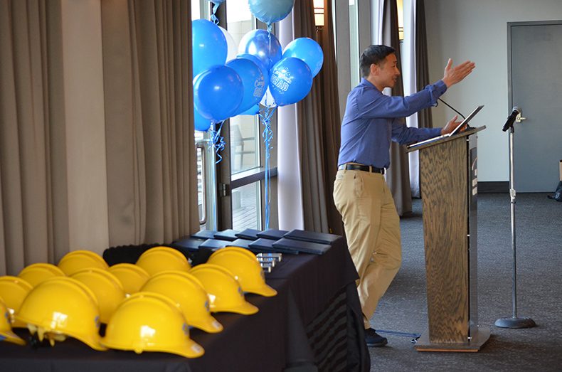 SPU General Manager and CEO Andrew Lee speaks at a podium, surrounded by blue graduation balloons and yellow SPU hardhats.