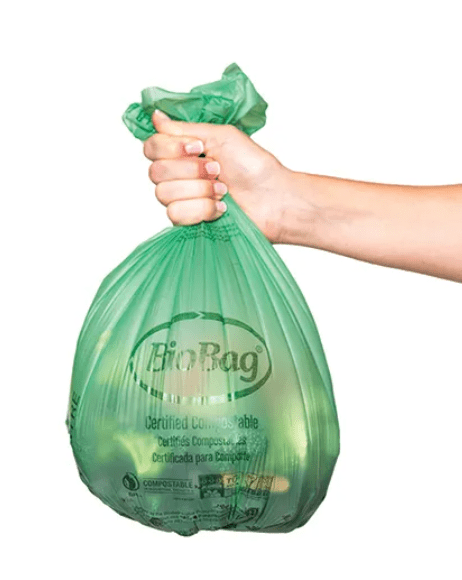 A photo that focuses on a full BioBag, this is a green compostable bag that is used to line kitchen compost bins for households to put their food scraps in until they’re ready to toss it in their compost cart. The bag is held at the top by an outstrechted arm form the right, the arm has a light skin tone.