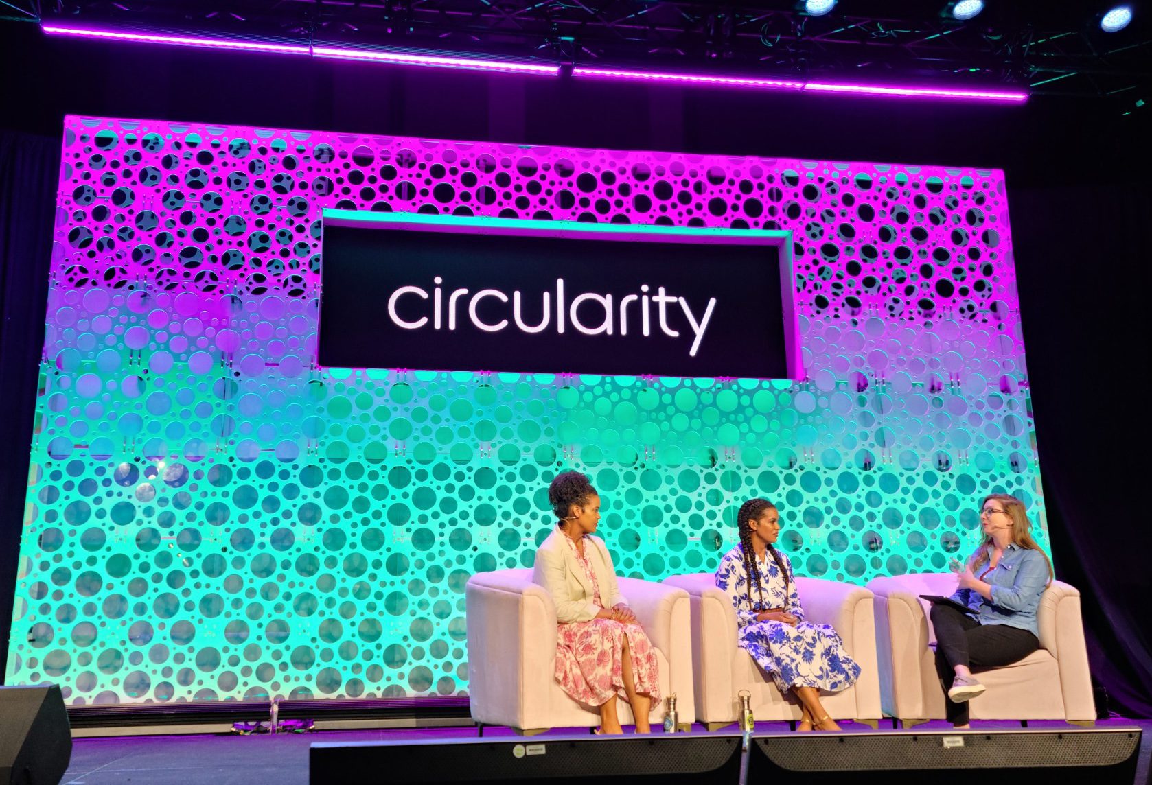 Three participants discussing issues on stage at Circularity 23, sitting on chairs in front of the brightly lit background.