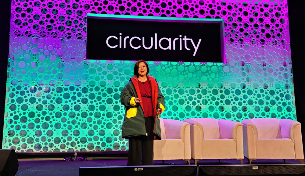 Ming-Ming Tung Edelman stands on stage at the Circularity Conference in front of a colorful, brightly lit, circle patterned backdrop.