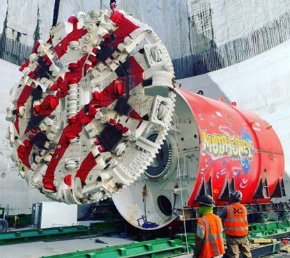 A large, round, red and white striped cutterhead is lowered onto the front of the Mudhoney tunnel-boring machine.