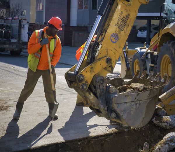 An SPU employee with shovel assists a backhoe digging a trench on city street.