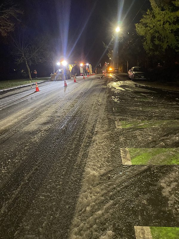 A road covered in snow and ice and lined with orange traffic cones with a work crew with a bulldozer working under bright work lights at night.