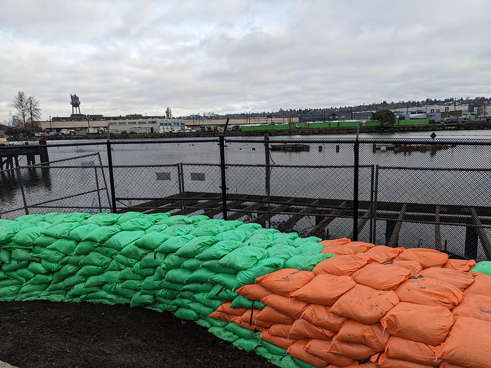 A wall of green and orange sandbags lines a fence next to a river.