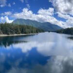 Blue sky and white clouds reflect off the surface of an alpine lake surrounded by mountains and evergreen forests.