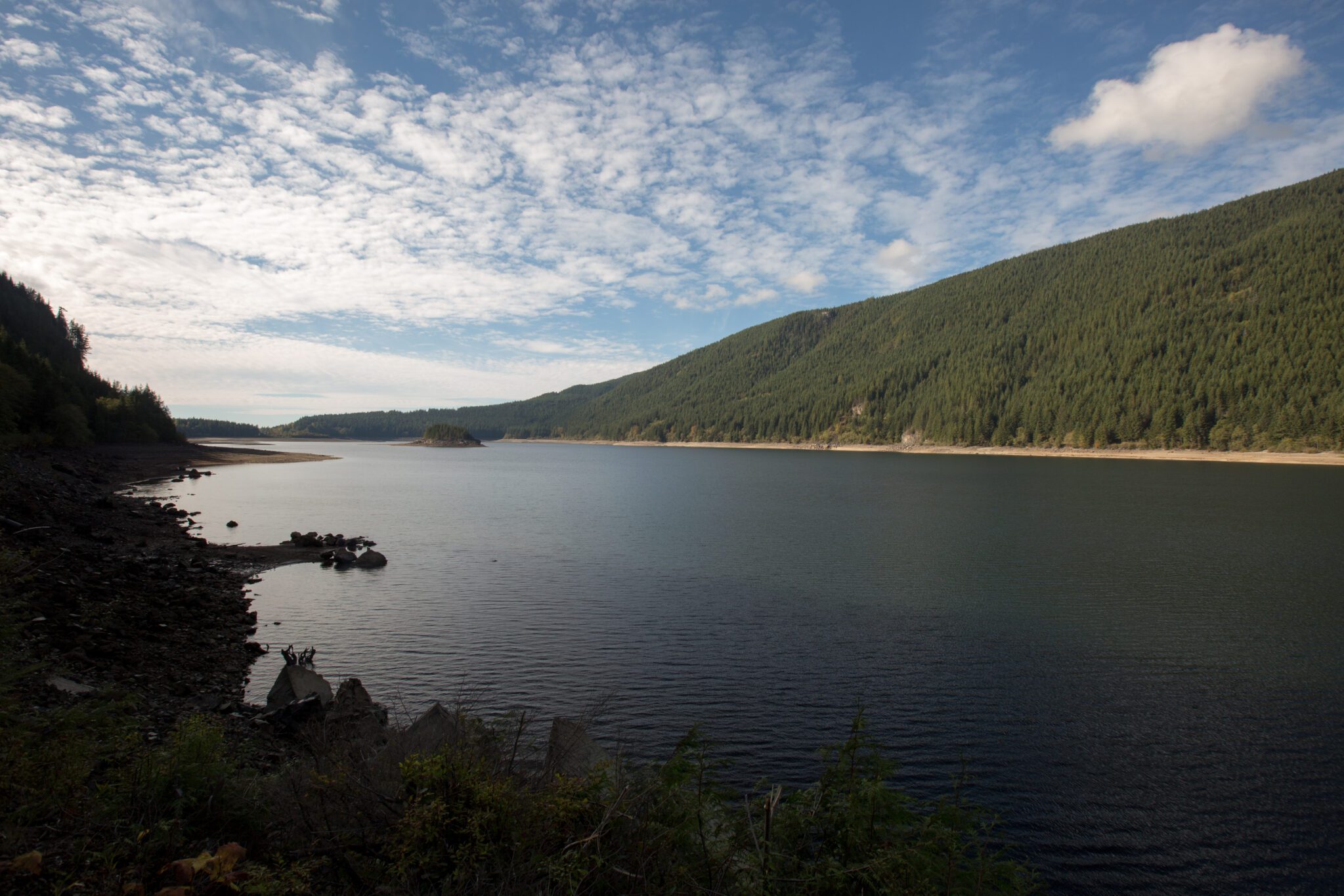 A lake surrounded by hills covered with evergreen forest under a blue sky dappled with white clouds.
