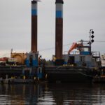 A barge on a river surrounded by a tug boat, cranes, and heavy industrial equipment.