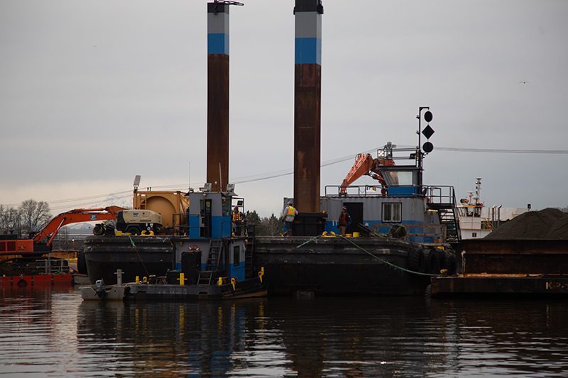 A barge on a river surrounded by a tug boat, cranes, and heavy industrial equipment.