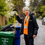 A person wearing an orange shirt, a dark jacket, and an ID badge stands on a residential street in light rain while measuring the height of items sticking out of a black waste bin.