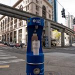 A cylindrical, blue water filling station with a water tumbler on it on a street corner in Seattle with an old stone building and monorail tracks behind it.