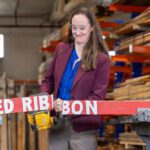 A woman in a lumber warehouse cuts through a red ribbon.