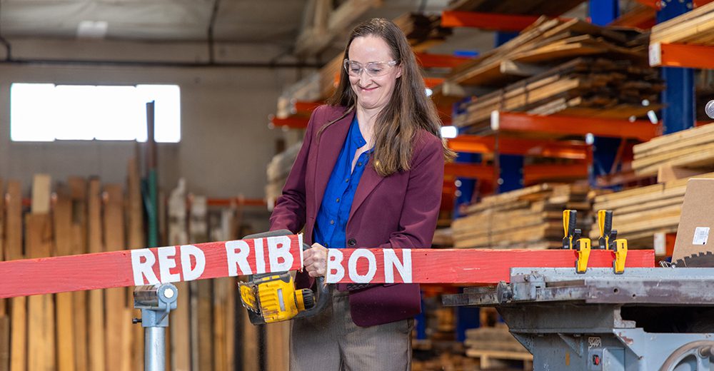 A woman in a lumber warehouse cuts through a red ribbon.