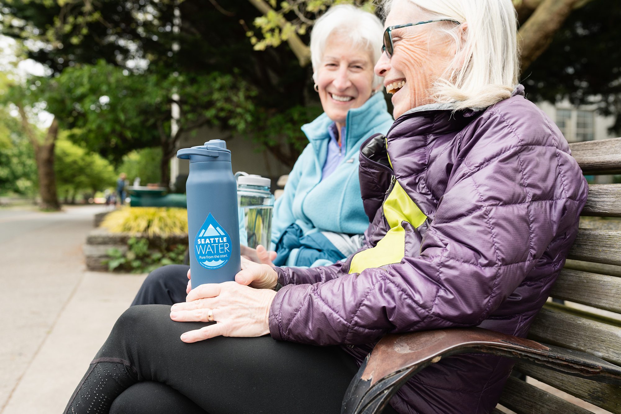 Two smiling women sit on a bench holding reusable water bottles with the "Seattle Water—Pure from the Start" logo.