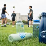 5 people kick a soccer ball on a grass field behind several water bottles with the Seattle Water—Pure from the Start logo.