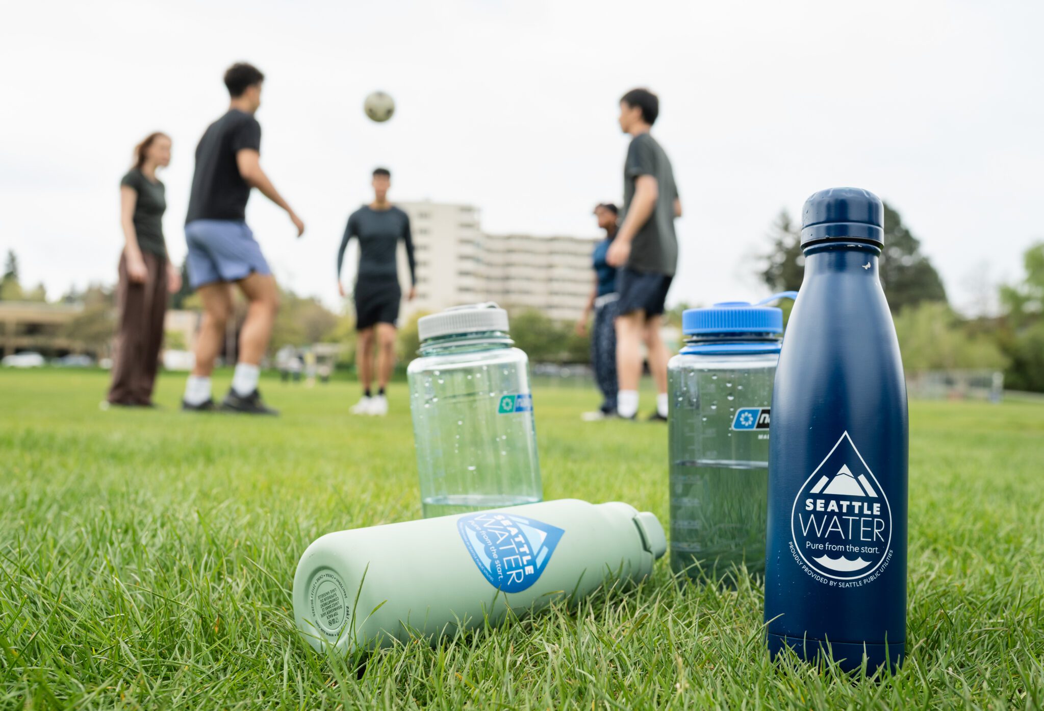 5 people kick a soccer ball on a grass field behind several water bottles with the Seattle Water—Pure from the Start logo.