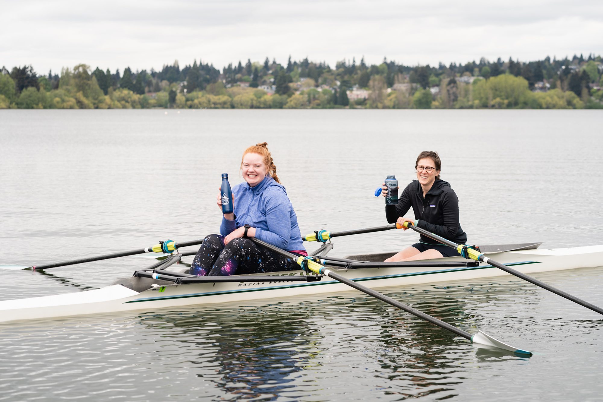 Two women in a kayak on a lake smile holding reusable water bottles with the "Seattle Water—Pure from the Start" logo.