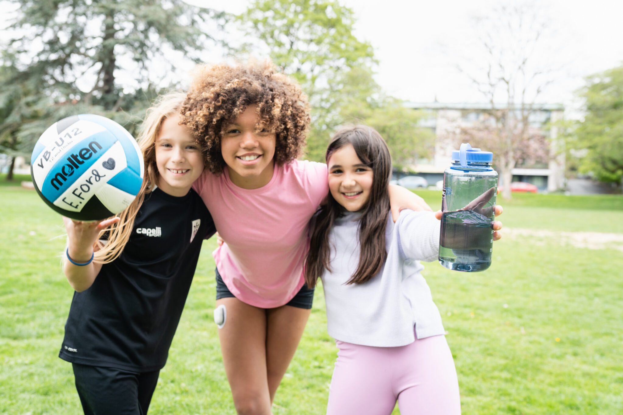 Three smiling girls pose with a soccer ball and a reusable water bottle on a grassy field.