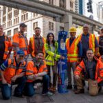 A group of a dozen or so SPU frontline employees in orange safety vests and hardhats pose next to a blue, SPU-branded water refill station in downtown Seattle under the Monorail tracks.