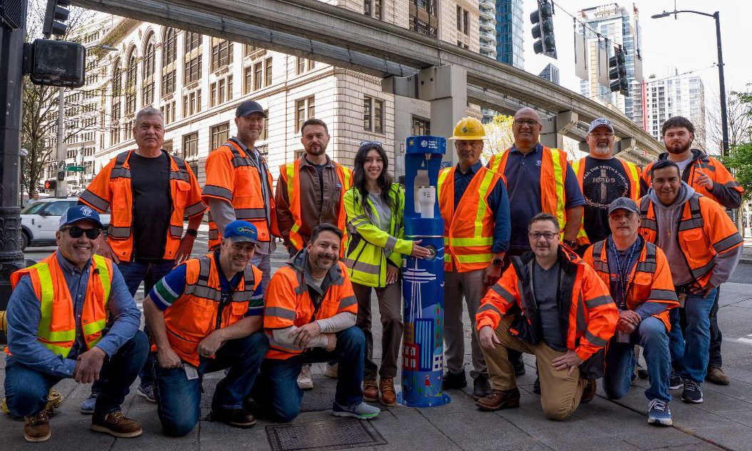 A group of a dozen or so SPU frontline employees in orange safety vests and hardhats pose next to a blue, SPU-branded water refill station in downtown Seattle under the Monorail tracks.